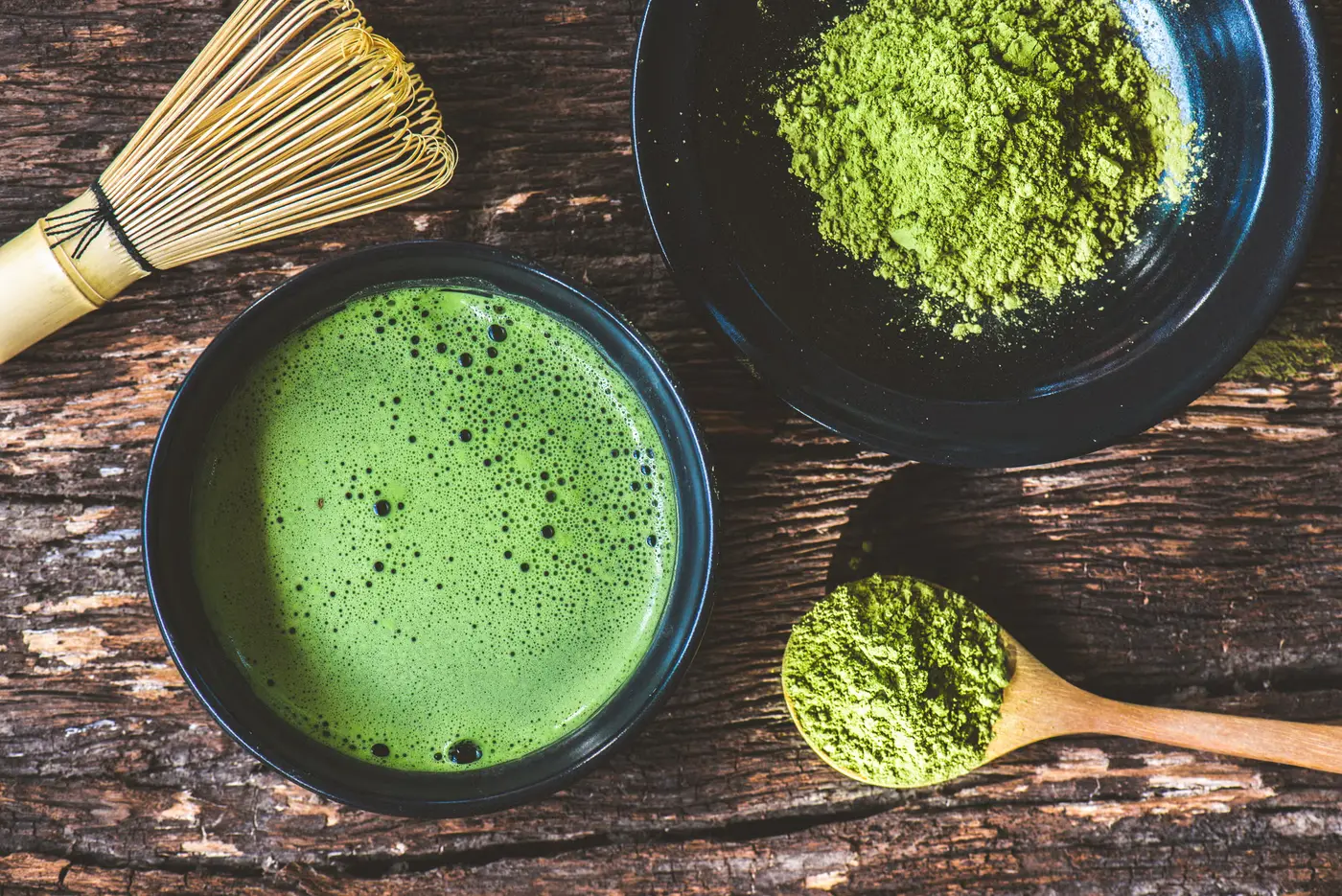 Overhead view of a bowl of whisked matcha beside a bamboo whisk, a dish of matcha powder and a wooden spoon on a dark wooden table